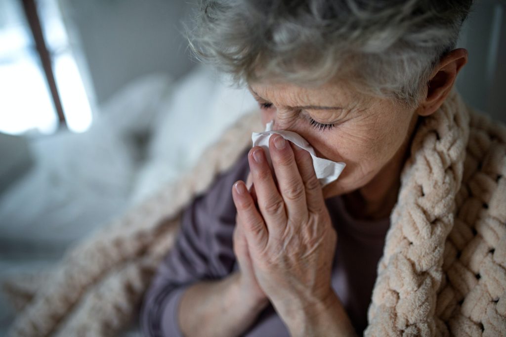 A mature woman wrapped in a blanket is blowing her nose with a tissue.