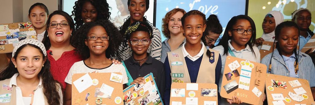 Three women stand with a group of school-aged children who are each holding up a cork board that has been decorated scrapbook-style.