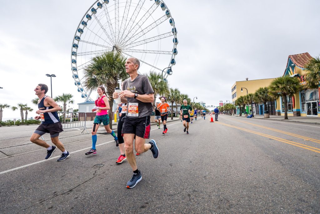 A group of people in activewear and running bibs run along a road with shops and a ferris wheel in the background.