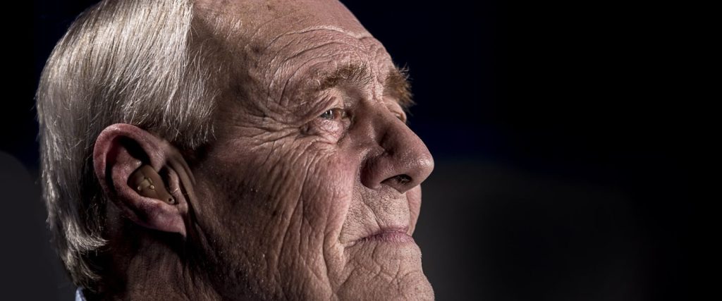 An older man with grey hair looks into the distance against a black background.