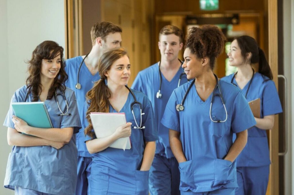 Four women and two men wearing blue scrubs with stethoscopes around their necks, walking along a corridor.