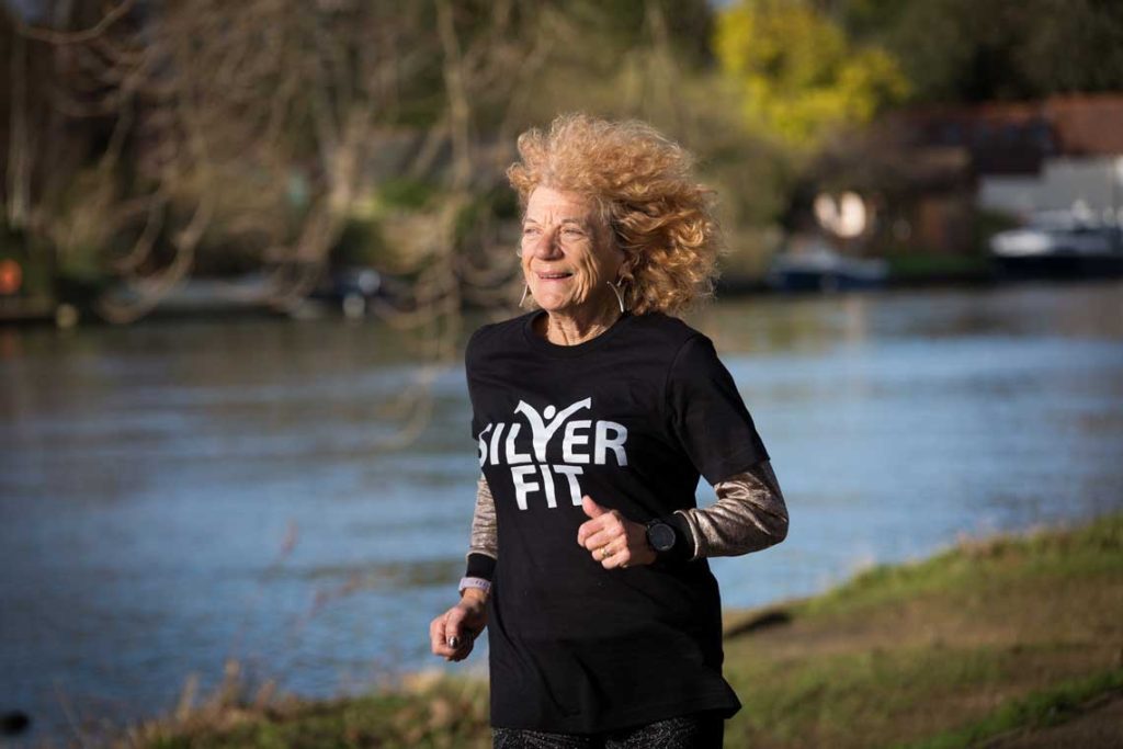 An older woman with strawberry blond hair runs along a riverbank, smiling, wearing a black t-shirt with Silver Fit written on the front.
