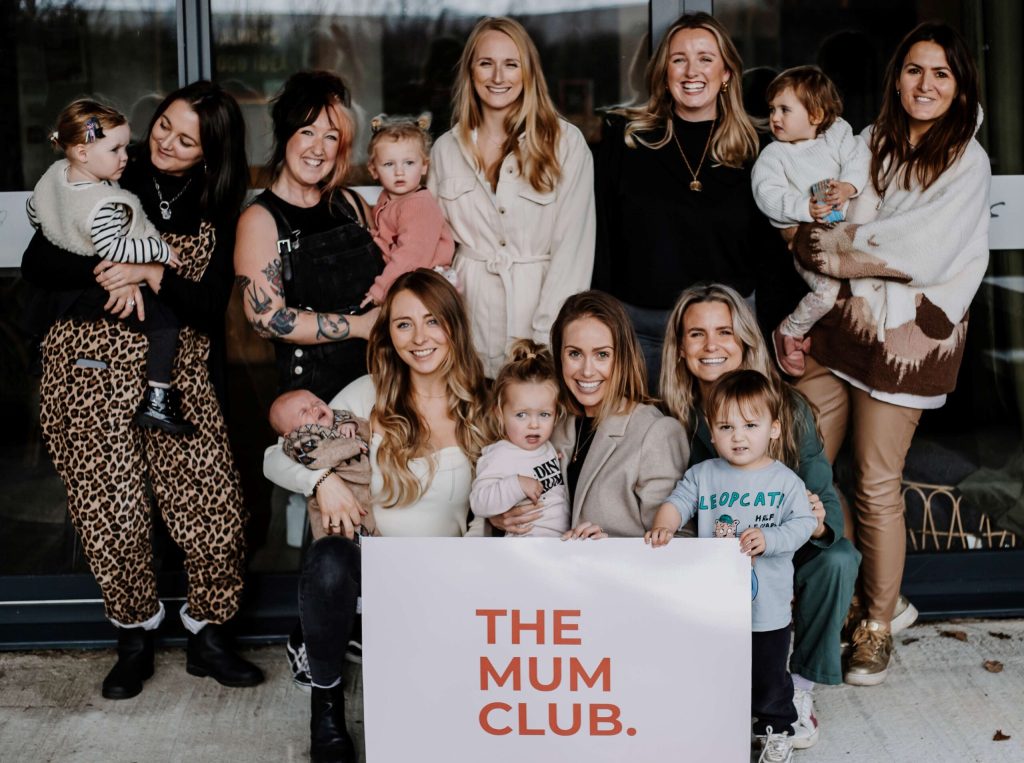 A group of mothers and their children pose in from of 'The Mum Club' sign.